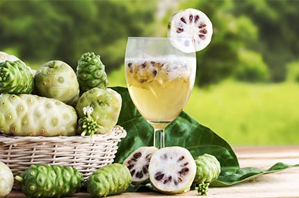 Glas mit Noni-Saft, umgeben von vielen gelblich-grünen Noni-Früchten